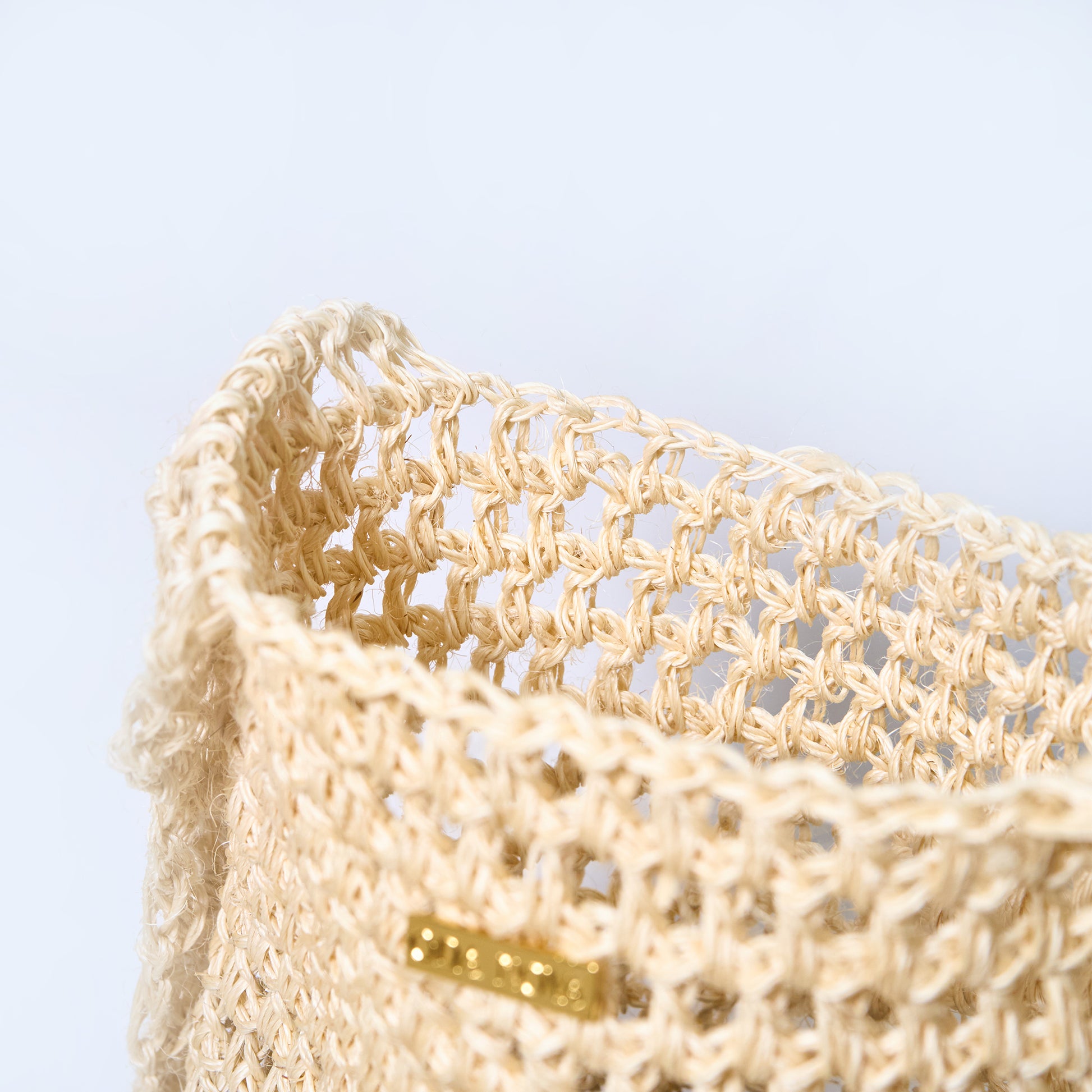 Beige woven basket with a gold tag on a light blue background. Aura cream crossbody woven bag handcrafted from natural fique fiber, designed by Oda Nova to honor feminine strength and Colombian craftsmanship.