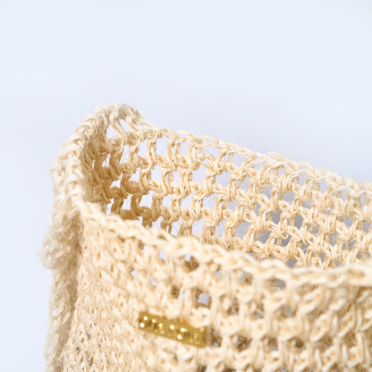 Beige woven basket with a gold tag on a light blue background. Aura cream crossbody woven bag handcrafted from natural fique fiber, designed by Oda Nova to honor feminine strength and Colombian craftsmanship.