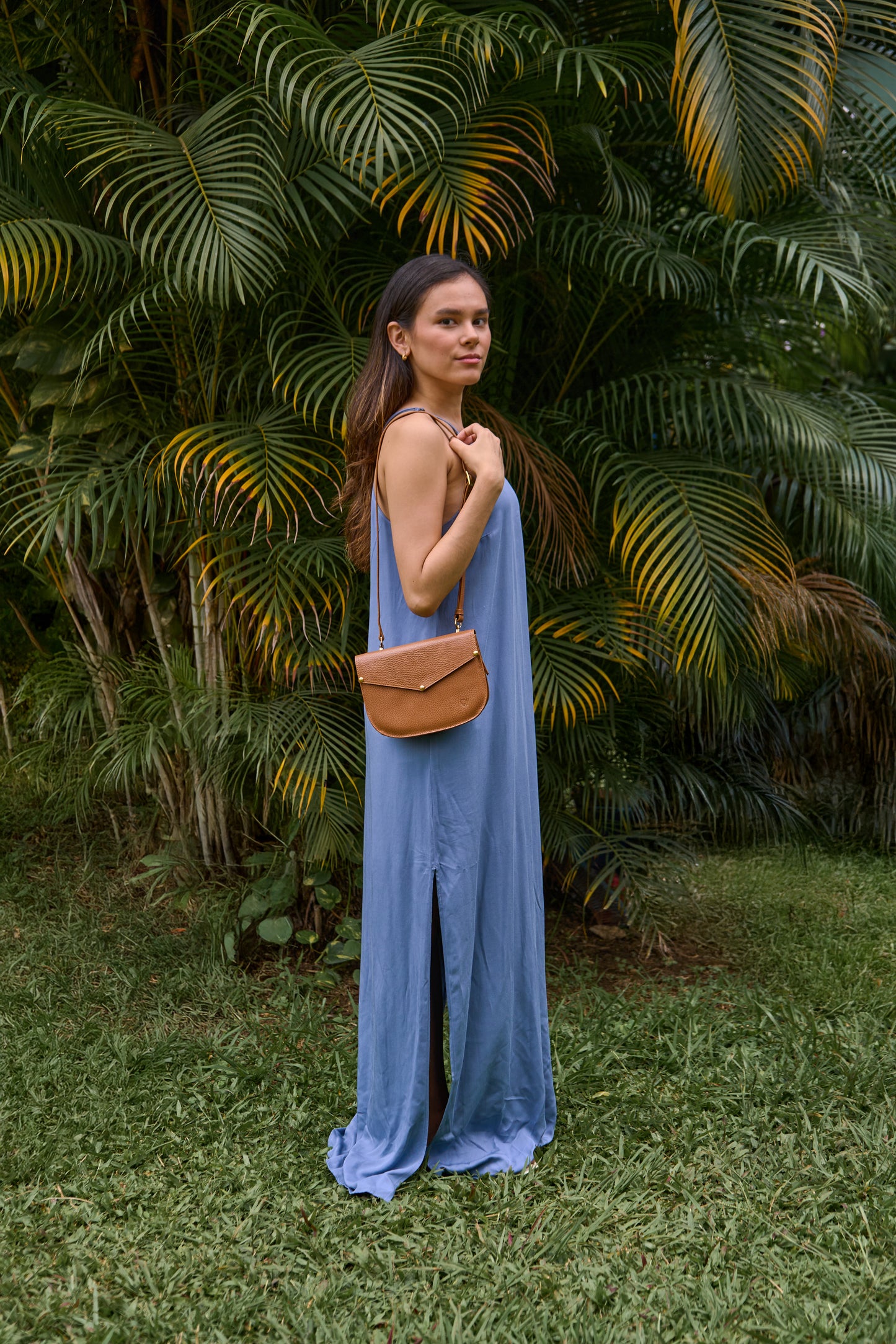 Model wearing Lyra leather crossbody bag a with a blue dress against a green leafy background