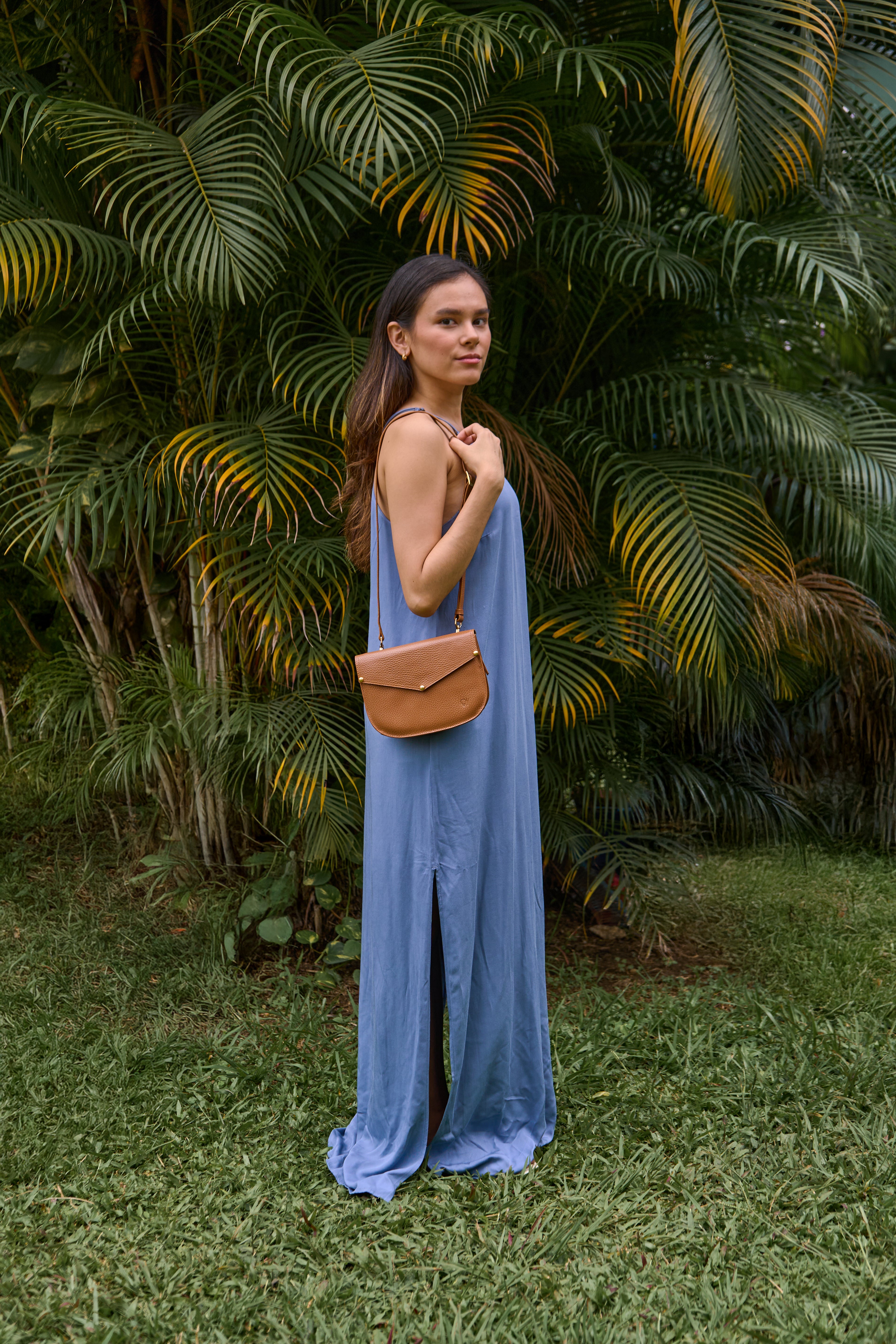 Model wearing Lyra leather crossbody bag a with a blue dress against a green leafy background