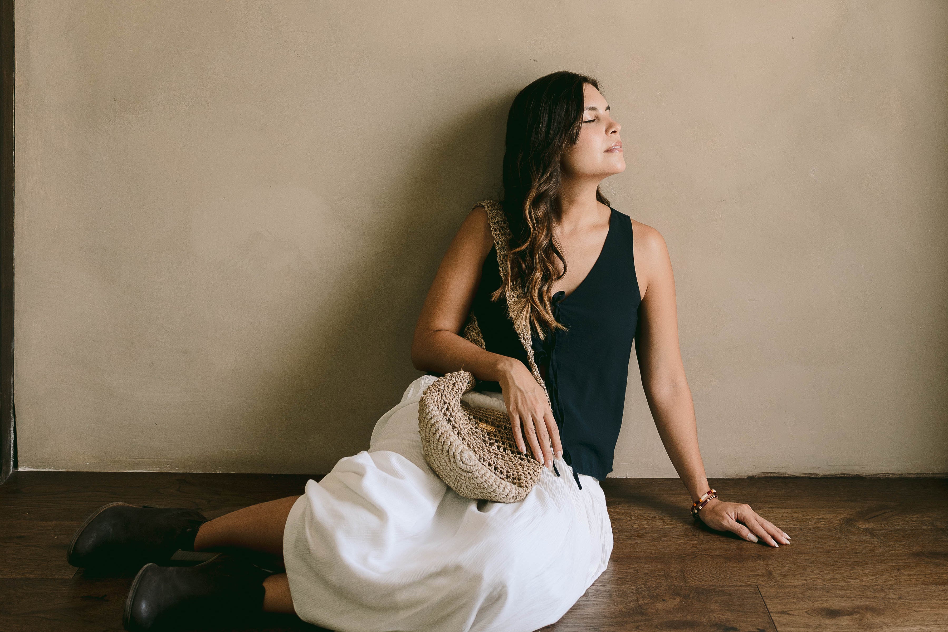 Woman sitting on the floor against a beige wall with a woven bag.