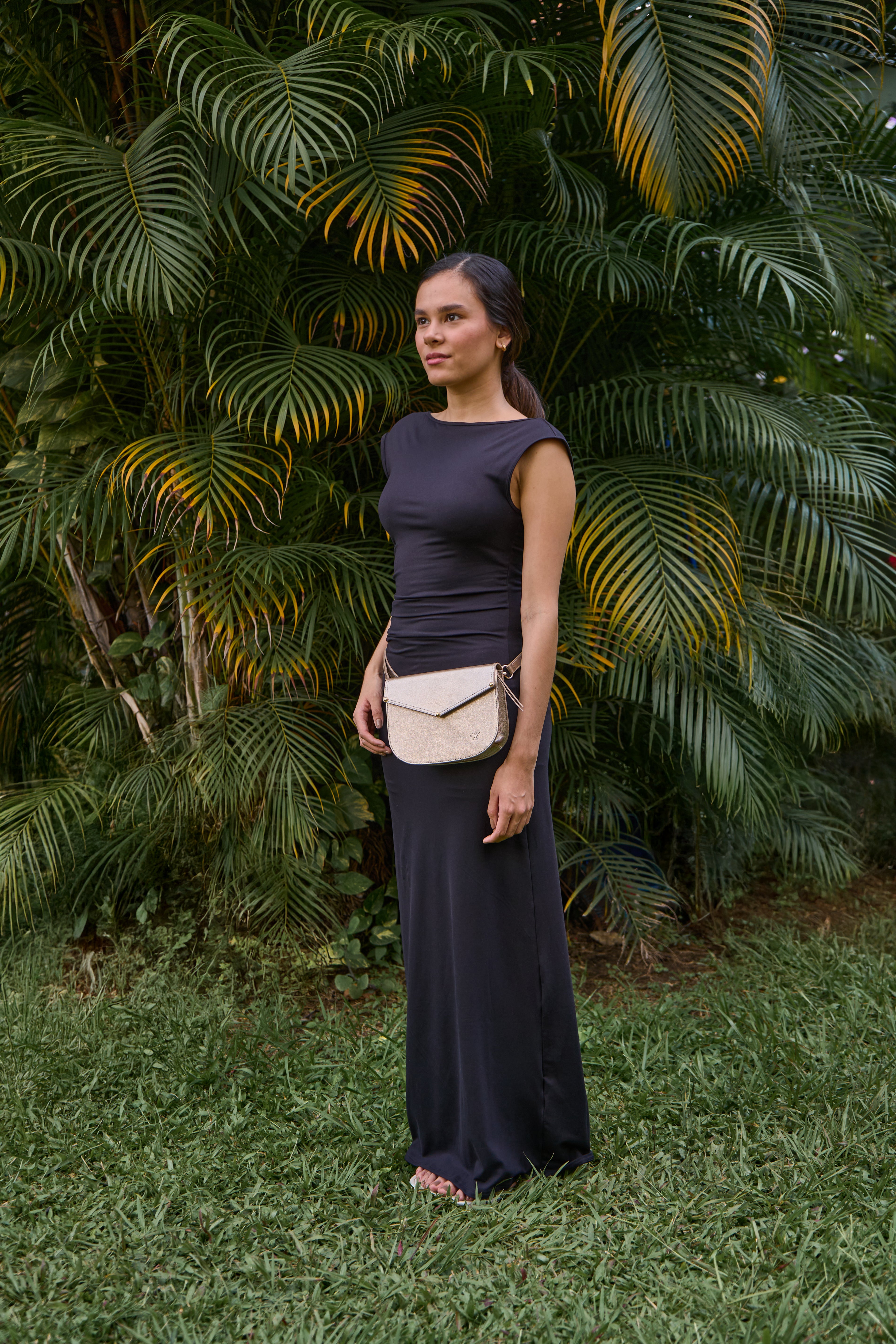 Woman in a dark dress standing in front of green foliage with bag across the waist